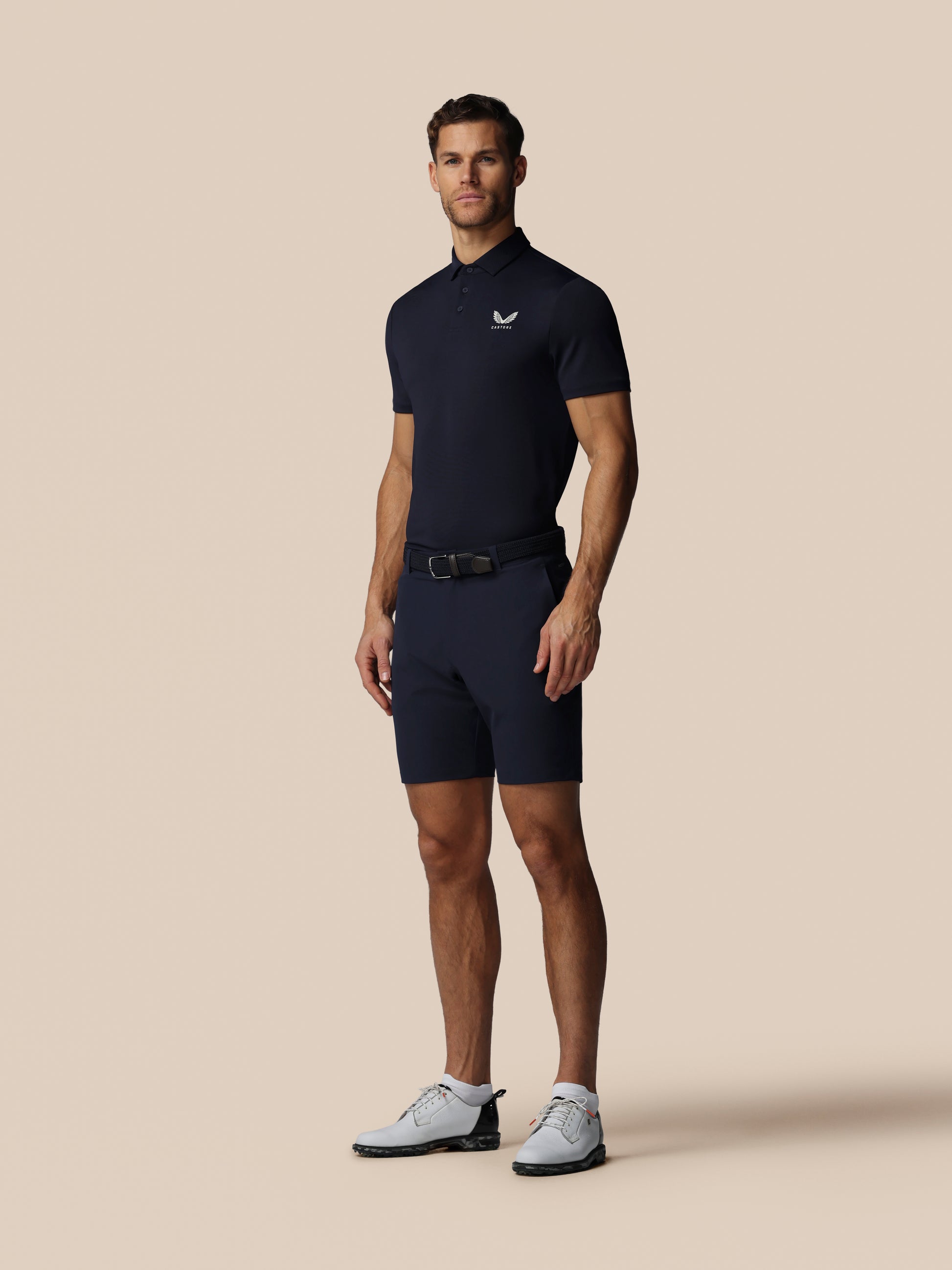 Man wearing a navy blue polo shirt and shorts with a belt on a beige background