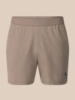 A pair of women's athletic shorts in a solid taupe color, featuring a wide elastic waistband with subtle branding and a small dark logo on the lower left leg, shown against a solid beige background.