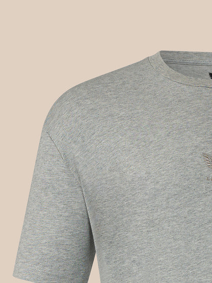 Close-up of a light heather gray crewneck t-shirt showing the shoulder, sleeve, and chest area against a solid beige background.