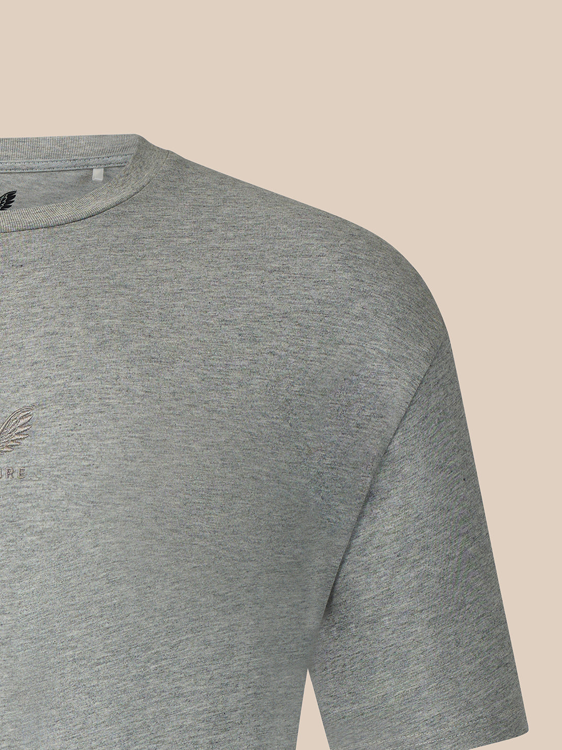 Close-up of a heather gray short-sleeve t-shirt showing the shoulder, collar, and a small tonal embroidered logo on the chest, set against a solid beige background.