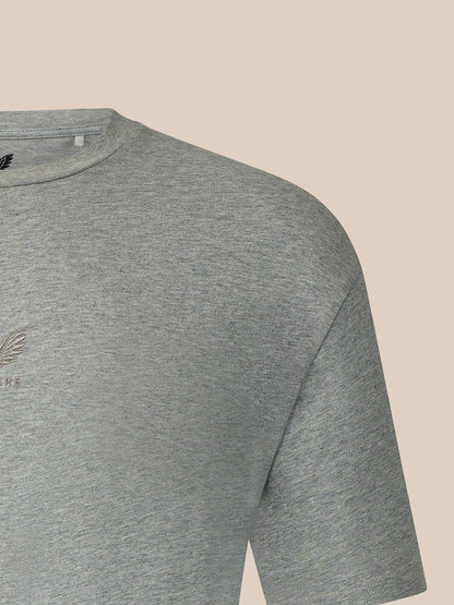 Close-up of a heather gray short-sleeve t-shirt showing the shoulder, collar, and a small tonal embroidered logo on the chest, set against a solid beige background.