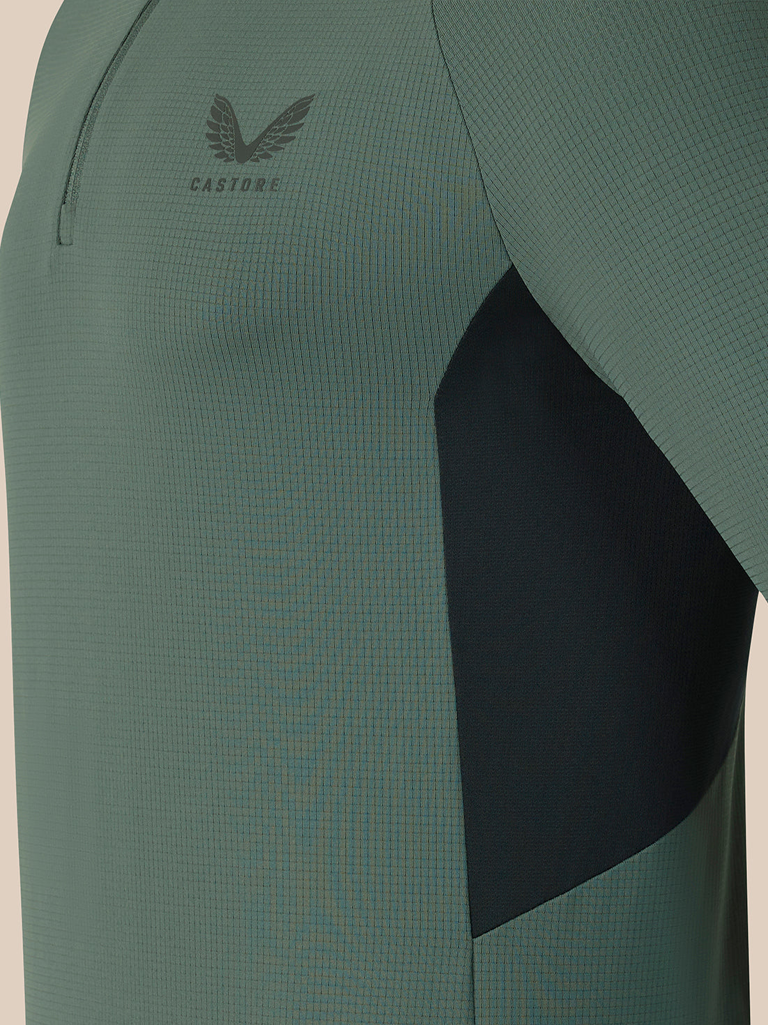Close-up of a Castore athletic top in sage green with a dark charcoal geometric side panel and a subtle wing logo on the chest.