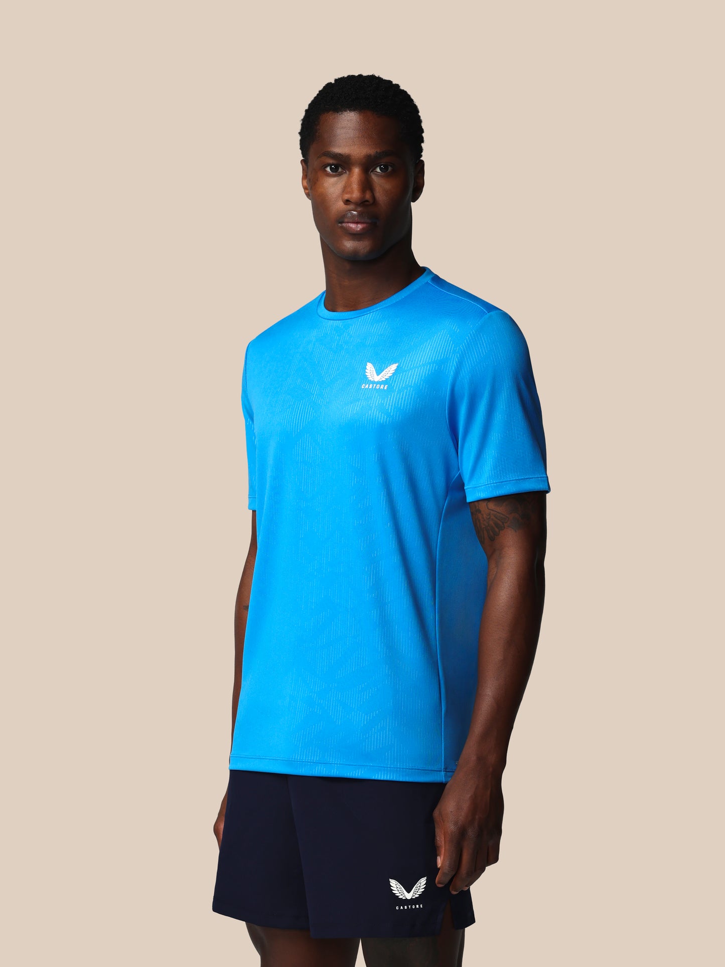 Man wearing a blue athletic shirt and black shorts with a logo on a beige background