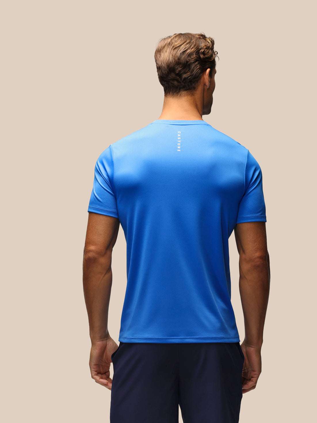 Back view of a man in a blue athletic short sleeve T-shirt with a vertical Castore logo on the upper back.