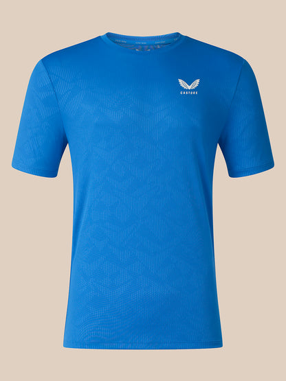 A blue short-sleeve athletic t-shirt with a subtle textured pattern and a small white winged logo on the left chest, displayed against a solid beige background.
