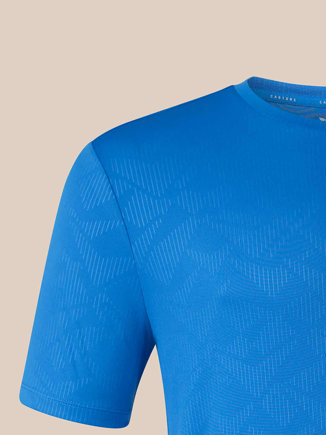 A close-up of a blue short-sleeve athletic t-shirt with a subtle geometric pattern, shown against a solid beige background.