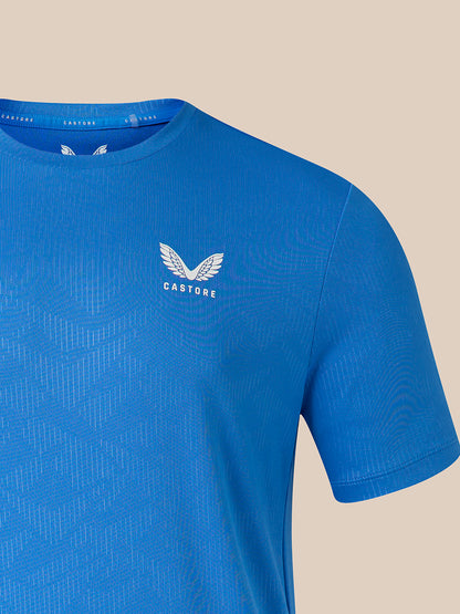 A close-up of a blue short-sleeve athletic t-shirt with a subtle geometric pattern, featuring a white winged logo and the brand name 'CASTORE' on the left chest, set against a solid beige background.