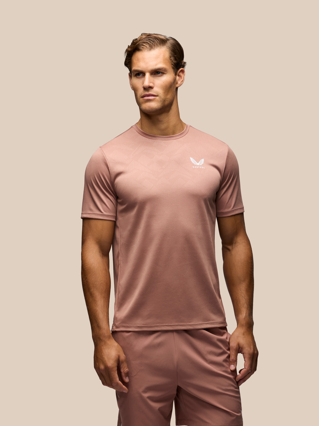 A man modeling a dusty rose athletic t-shirt and matching shorts against a solid beige background. The shirt features a small white wing logo on the left chest.