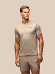 A male model wearing a light taupe short-sleeve athletic t-shirt and matching shorts against a solid light beige background.