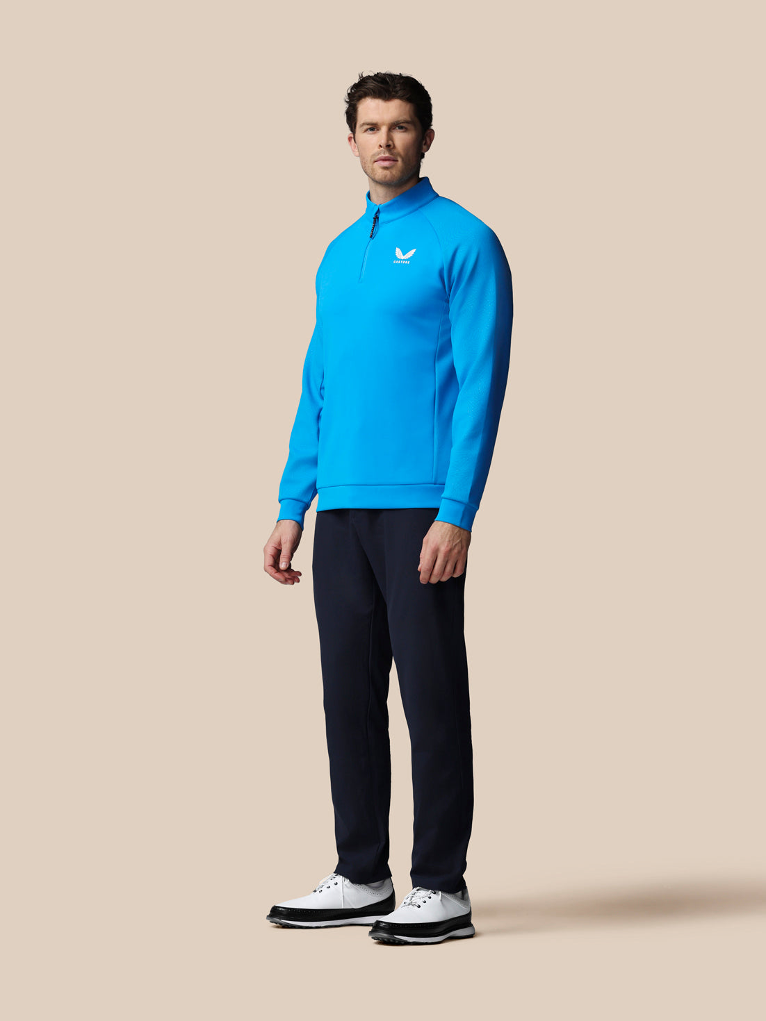 Man wearing a blue quarter zip top and navy golf trousers on a beige background