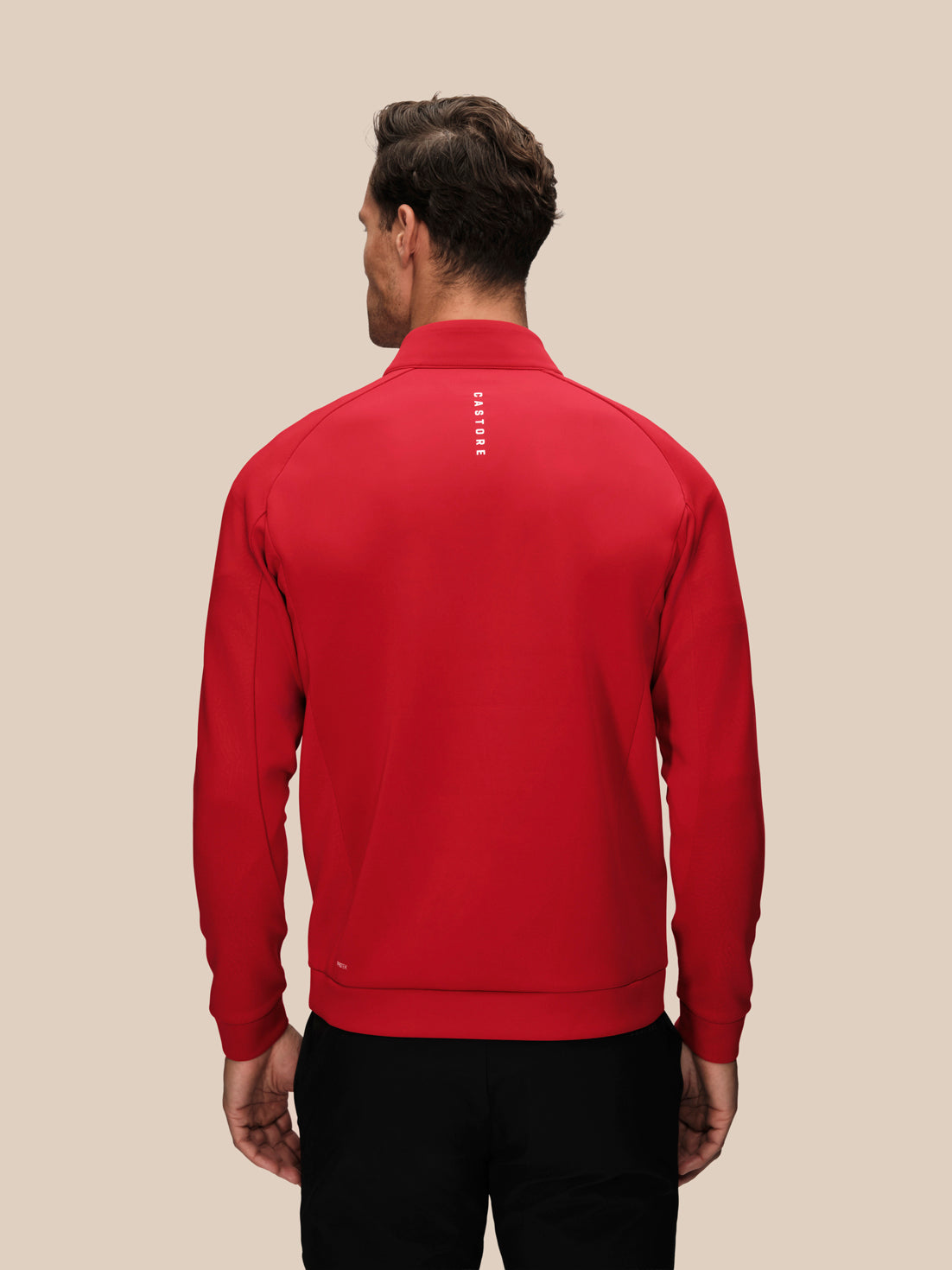 A man viewed from the back wearing a bright red long-sleeve quarter-zip pullover with a small vertical white logo on the upper back, paired with black pants, against a solid beige background.
