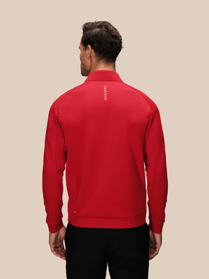 A man viewed from the back wearing a bright red long-sleeve quarter-zip pullover with a small vertical white logo on the upper back, paired with black pants, against a solid beige background.