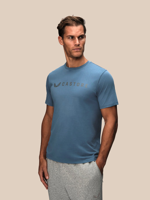 A man wearing a blue Castore athletic t-shirt and gray sweatpants, standing against a solid beige background.