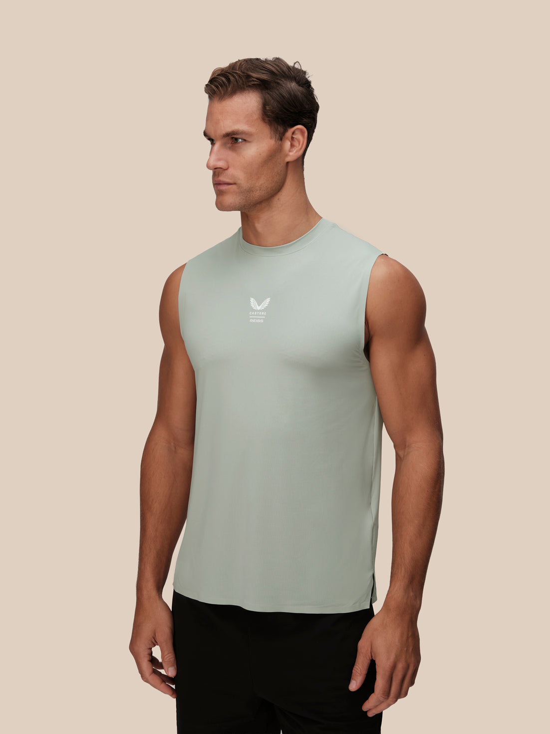 A muscular man wearing a sleeveless light sage green athletic tank top with a small white logo on the chest, paired with black pants, set against a solid beige background.