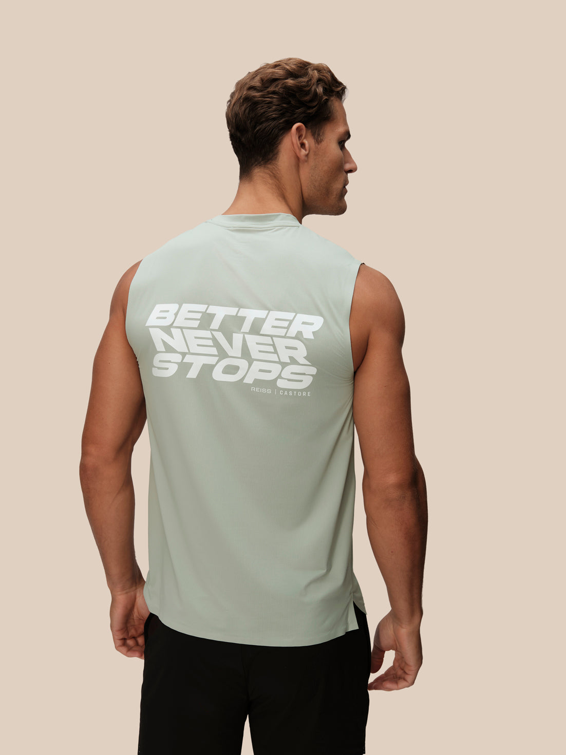 A man shown from the back wearing a sage green sleeveless athletic tank top with a large white graphic text print reading 'BETTER NEVER STOPS' on the back, paired with black athletic pants, against a solid beige background.