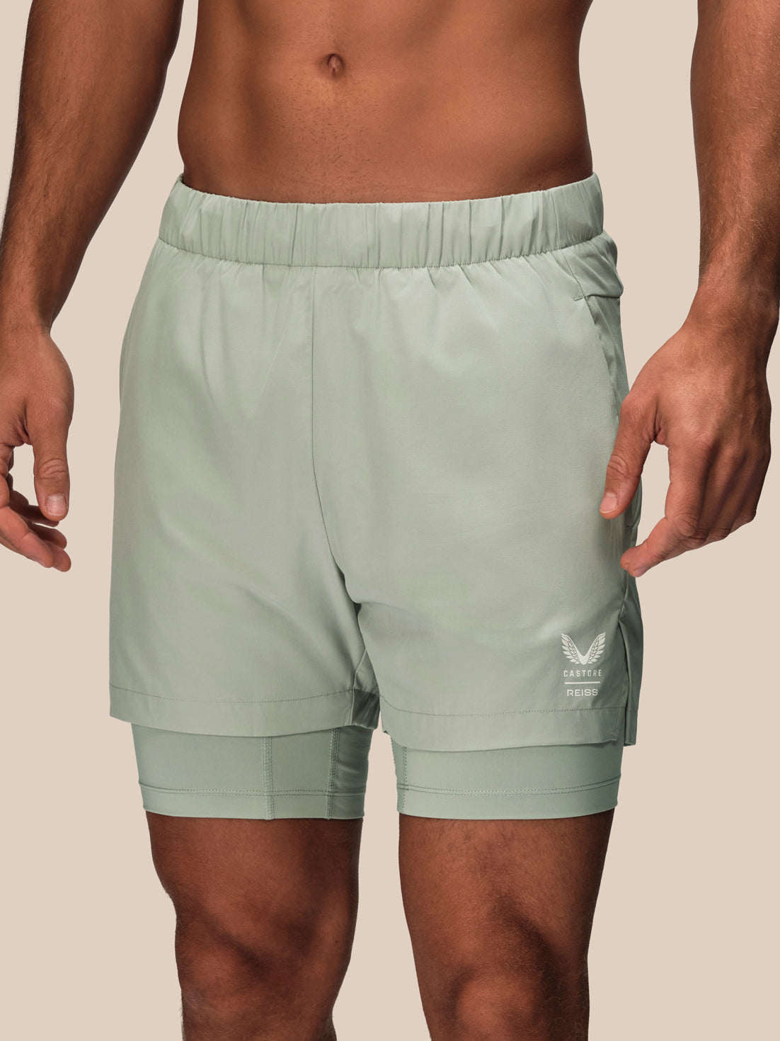 A close-up of a man wearing sage green double-layered athletic shorts with a white logo on the lower left leg, set against a solid beige background.