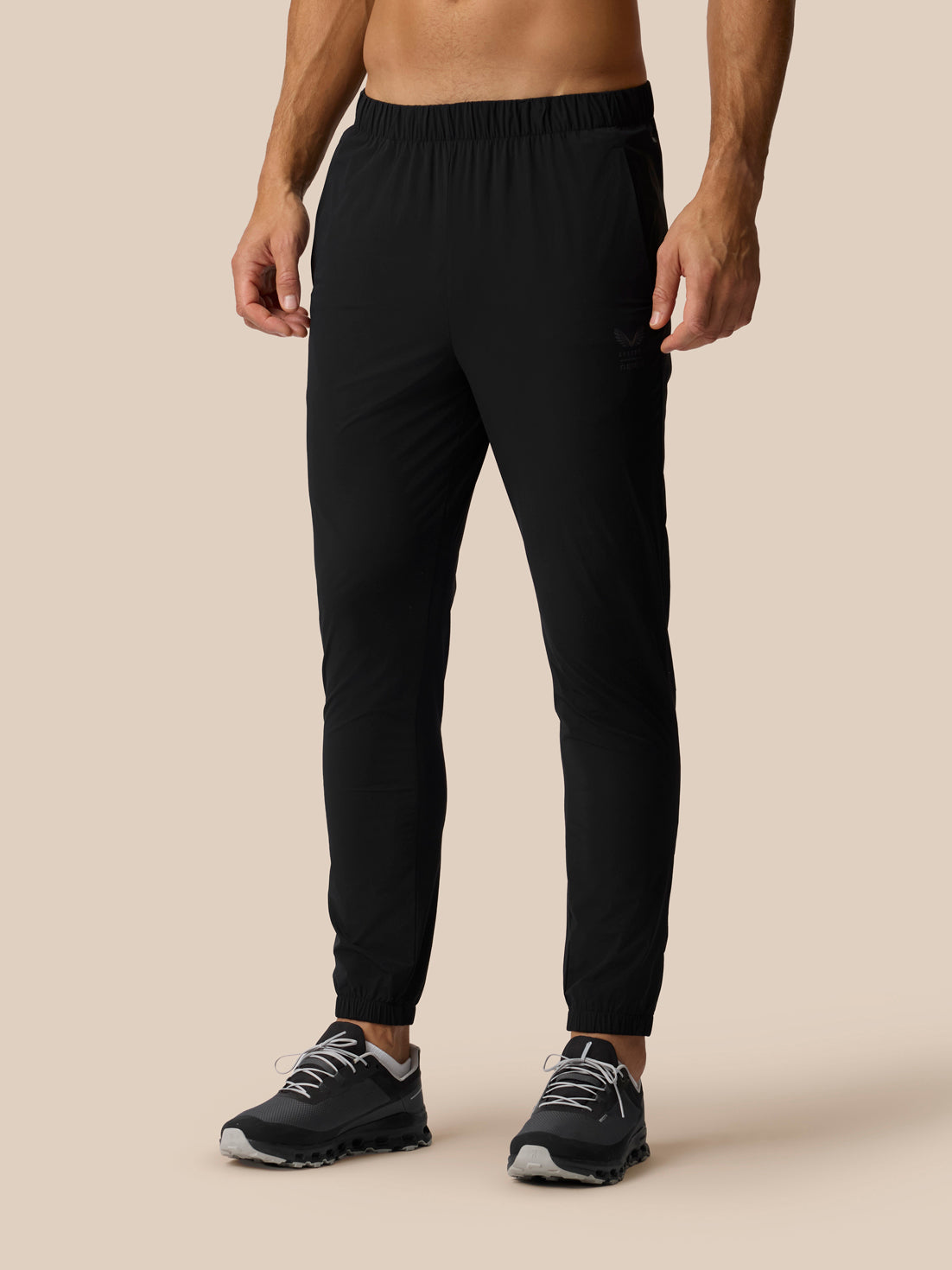 A man wearing black jogger-style athletic pants and black sneakers, shown from the waist down against a plain beige studio background.