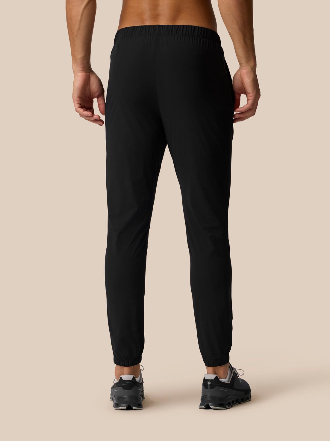 Rear view of a person wearing black jogger-style pants and dark sneakers against a plain beige studio background.
