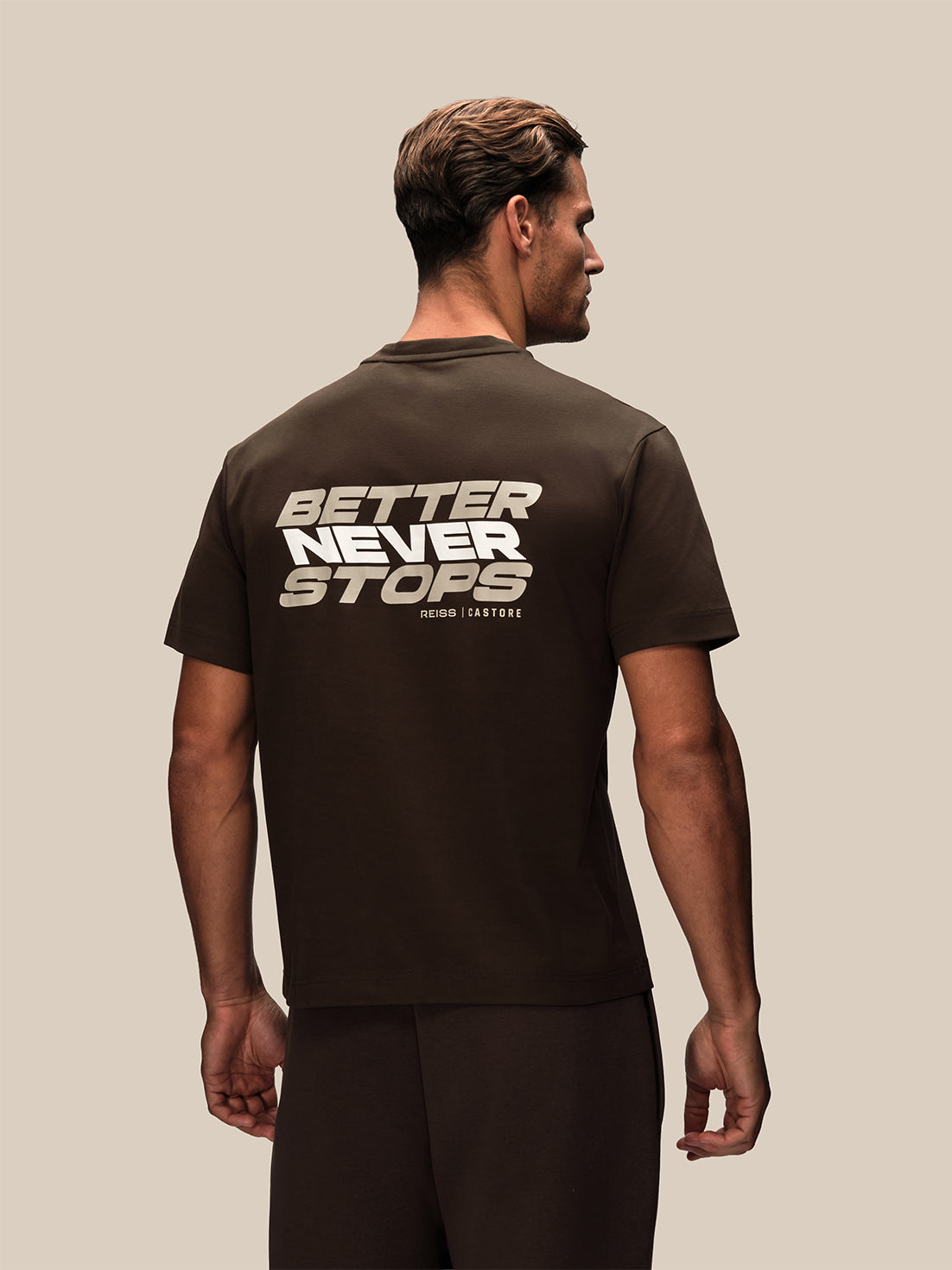 A man shown from the back wearing a dark brown t-shirt with a large graphic print on the back reading 'BETTER NEVER STOPS' in white and beige, paired with dark brown pants, against a solid beige background.