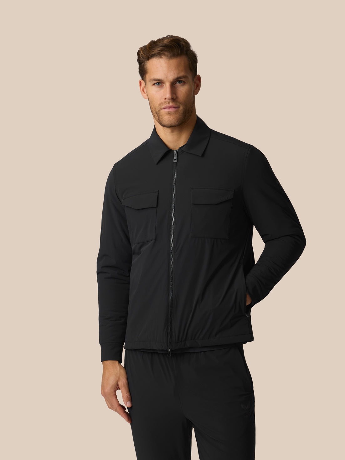 A man wearing a black zip-front jacket with a collar and two chest flap pockets, paired with black pants, photographed against a plain warm beige studio background.