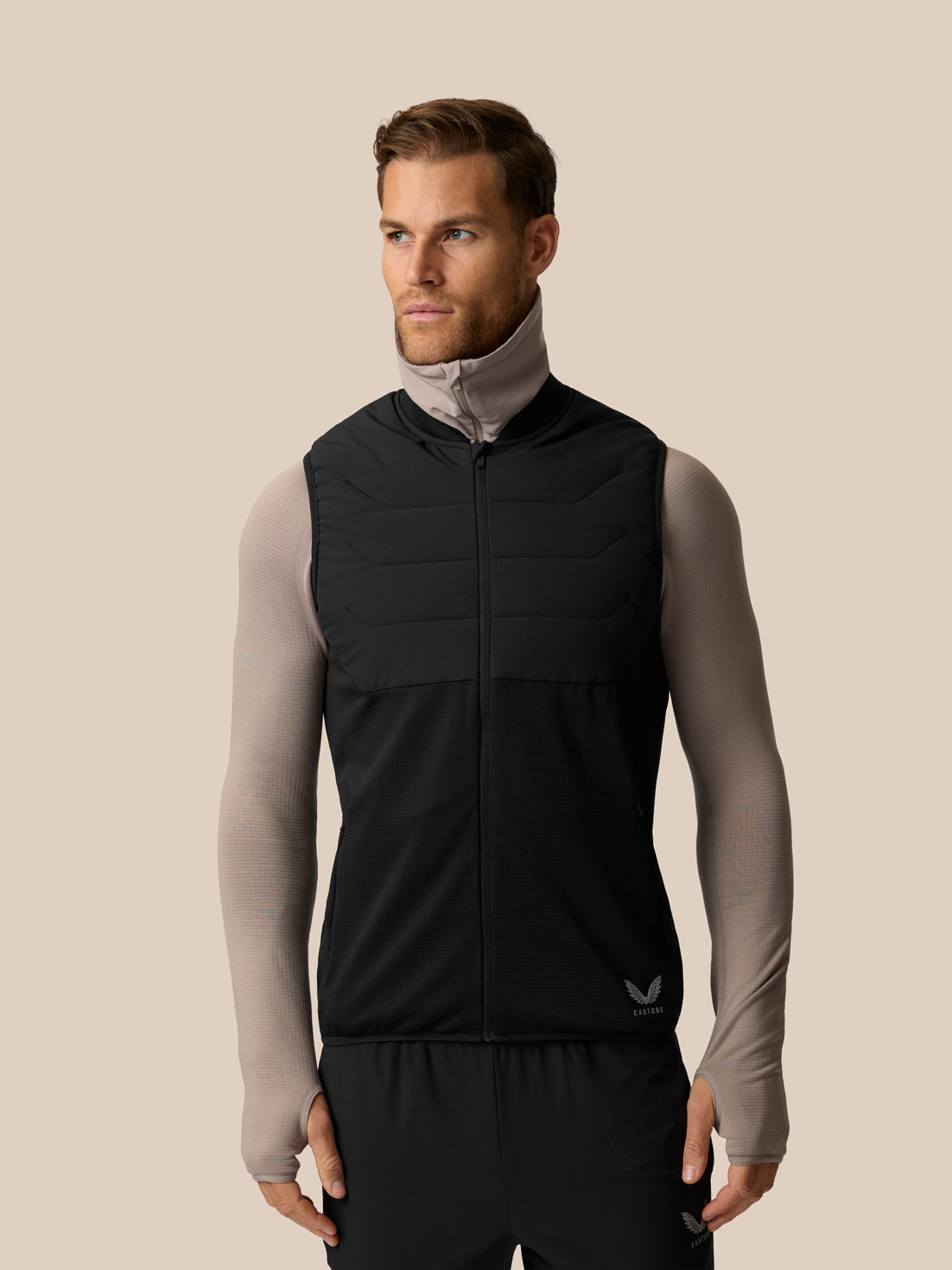 A man wearing a black sleeveless zip-front athletic vest over a light gray long-sleeve top, standing against a plain beige studio background.