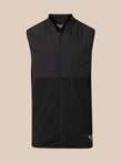 A black sleeveless zip-up vest with a mock neck and horizontal quilting, shown against a solid beige background. A small white logo is visible on the bottom right hem.