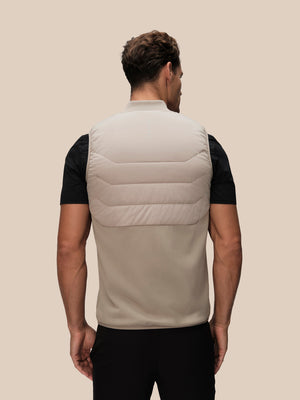 Back view of a taupe thermo hybrid gilet worn over a black T shirt showing quilted upper panels and a vertical logo at the center back.