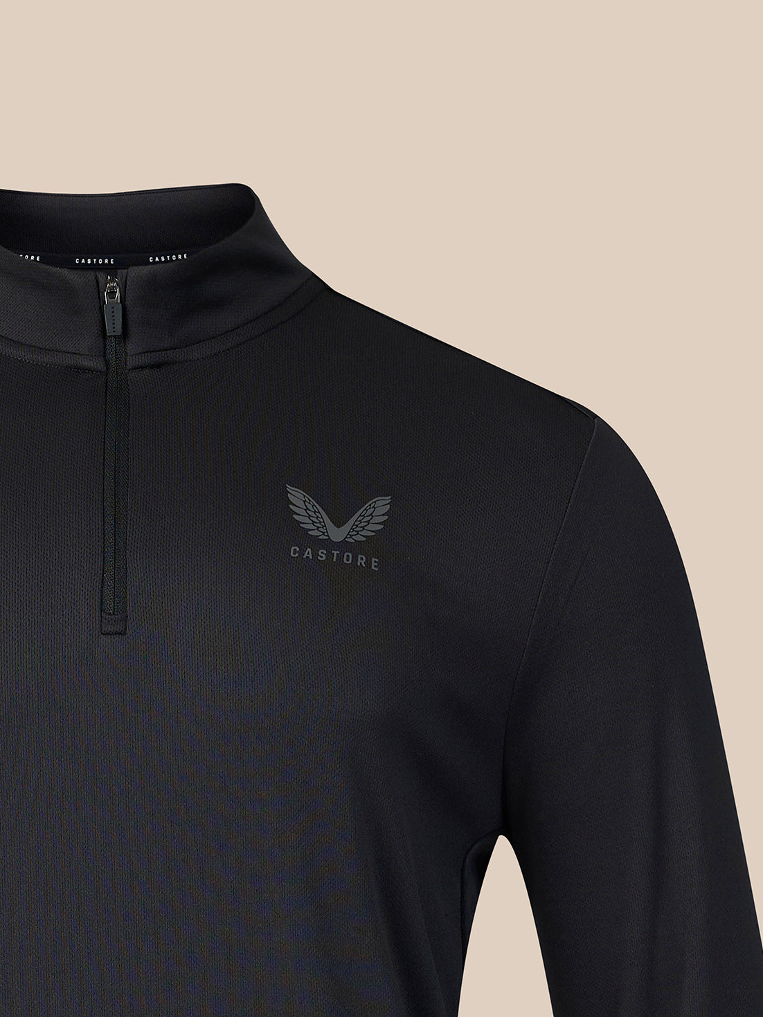 Close-up of a black long-sleeve quarter-zip pullover with a subtle gray winged logo and the word 'CASTORE' on the left chest, set against a solid beige background.