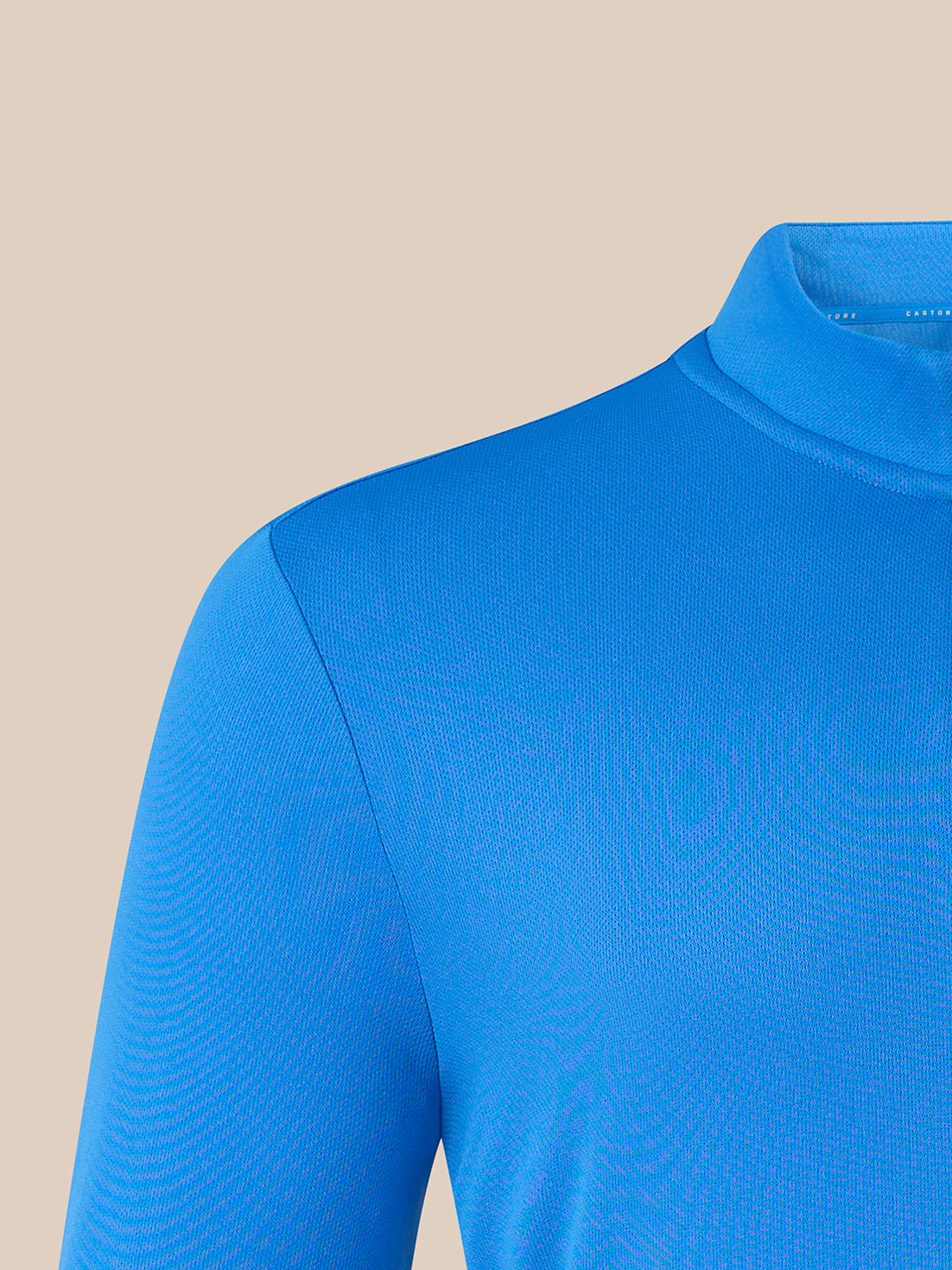 A close-up of a bright blue long-sleeve mock neck top, showing the ribbed texture of the fabric and the collar, set against a solid beige background.