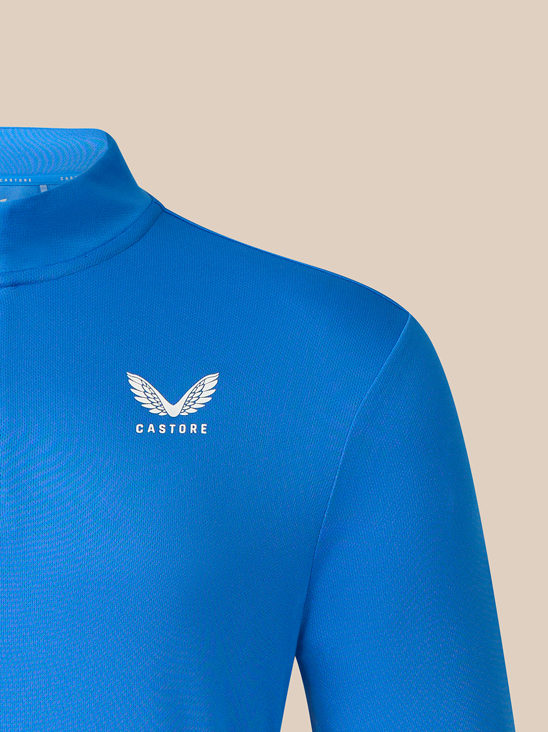A close-up of a bright blue long-sleeve athletic top with a mock neck collar, featuring a white Castore wing logo on the left chest, set against a solid beige background.