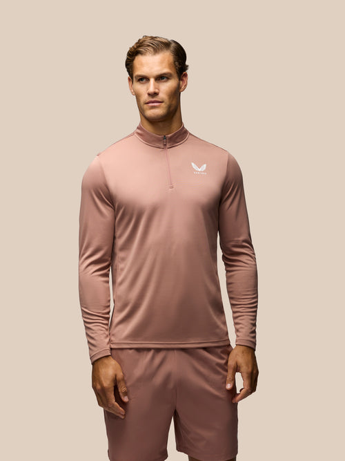 A man modeling a dusty rose long-sleeve athletic quarter-zip pullover with matching shorts against a solid beige background.
