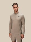 A man modeling a light taupe long-sleeve quarter-zip athletic top with a small dark logo on the left chest, paired with matching shorts, against a solid light beige background.
