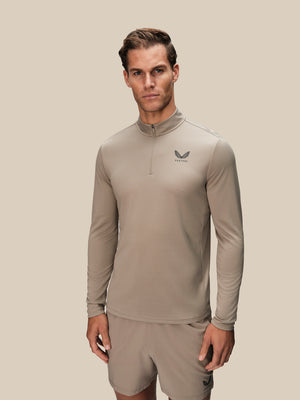 A man modeling a light taupe long-sleeve quarter-zip athletic top with a small dark logo on the left chest, paired with matching shorts, against a solid light beige background.