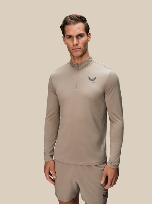 A man modeling a light taupe long-sleeve quarter-zip athletic top with a small dark logo on the left chest, paired with matching shorts, against a solid light beige background.
