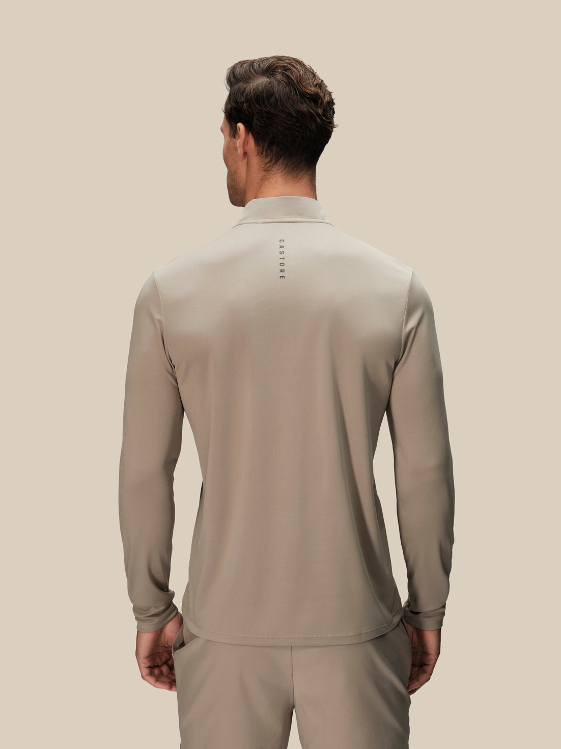 A man viewed from the back wearing a light taupe long-sleeve athletic quarter-zip pullover and matching pants against a solid beige background.