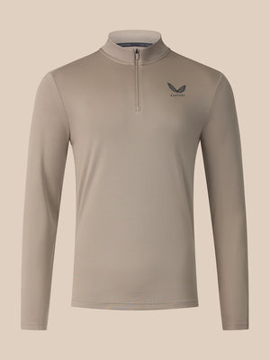 A long-sleeve quarter-zip athletic top in a light taupe-beige color, featuring a small dark gray wing logo and the brand name 'Castore on the left chest, displayed against a solid light beige background.