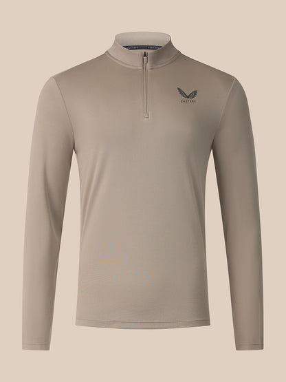 A long-sleeve quarter-zip athletic top in a light taupe-beige color, featuring a small dark gray wing logo and the brand name 'Castore on the left chest, displayed against a solid light beige background.