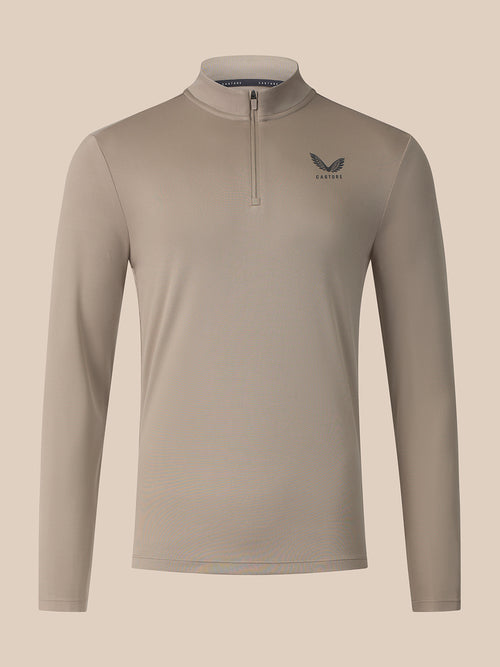 A long-sleeve quarter-zip athletic top in a light taupe-beige color, featuring a small dark gray wing logo and the brand name 'Castore on the left chest, displayed against a solid light beige background.