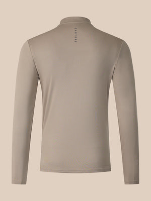 A long-sleeve athletic top shown from the back, featuring a stand-up collar and a small vertical 'CASTORE' logo printed in black near the center of the neck. The garment is a solid taupe color, displayed on a clean, solid, warm beige background.