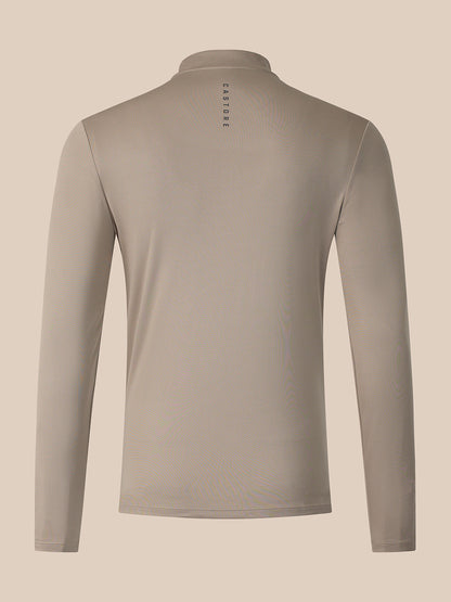 A long-sleeve athletic top shown from the back, featuring a stand-up collar and a small vertical 'CASTORE' logo printed in black near the center of the neck. The garment is a solid taupe color, displayed on a clean, solid, warm beige background.
