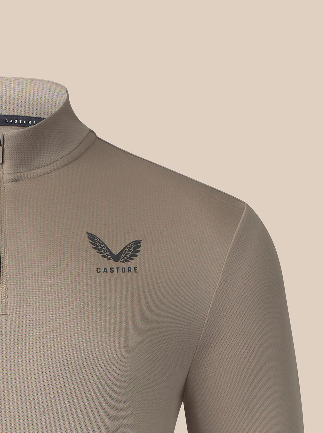 Close-up of a Castore quarter-zip neckwear garment in a warm taupe-beige color, featuring a dark gray winged logo and brand name on the chest.