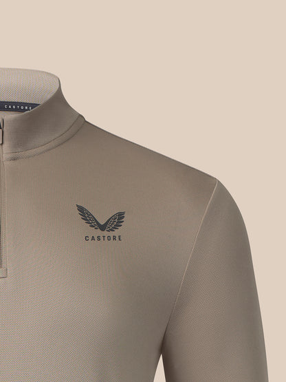 Close-up of a Castore quarter-zip neckwear garment in a warm taupe-beige color, featuring a dark gray winged logo and brand name on the chest.