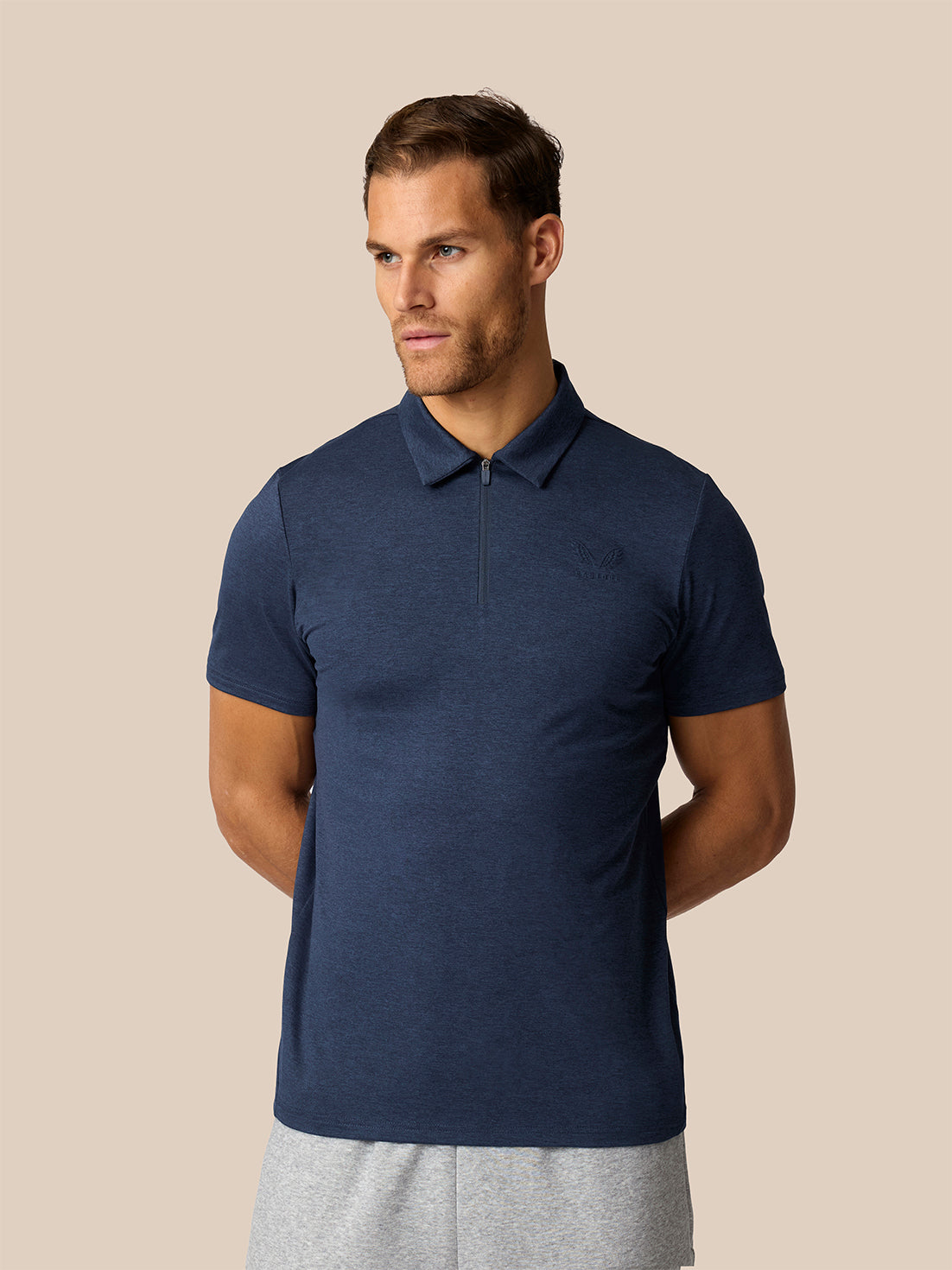 A man wearing a navy blue short-sleeve polo shirt with a half-zip collar, paired with light gray sweatpants, set against a solid beige background.