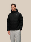 A man wearing a black hooded puffer jacket with a small silver logo on the left chest, paired with gray sweatpants, standing against a solid beige background.