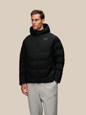A man wearing a black hooded puffer jacket with a small silver logo on the left chest, paired with gray sweatpants, standing against a solid beige background.