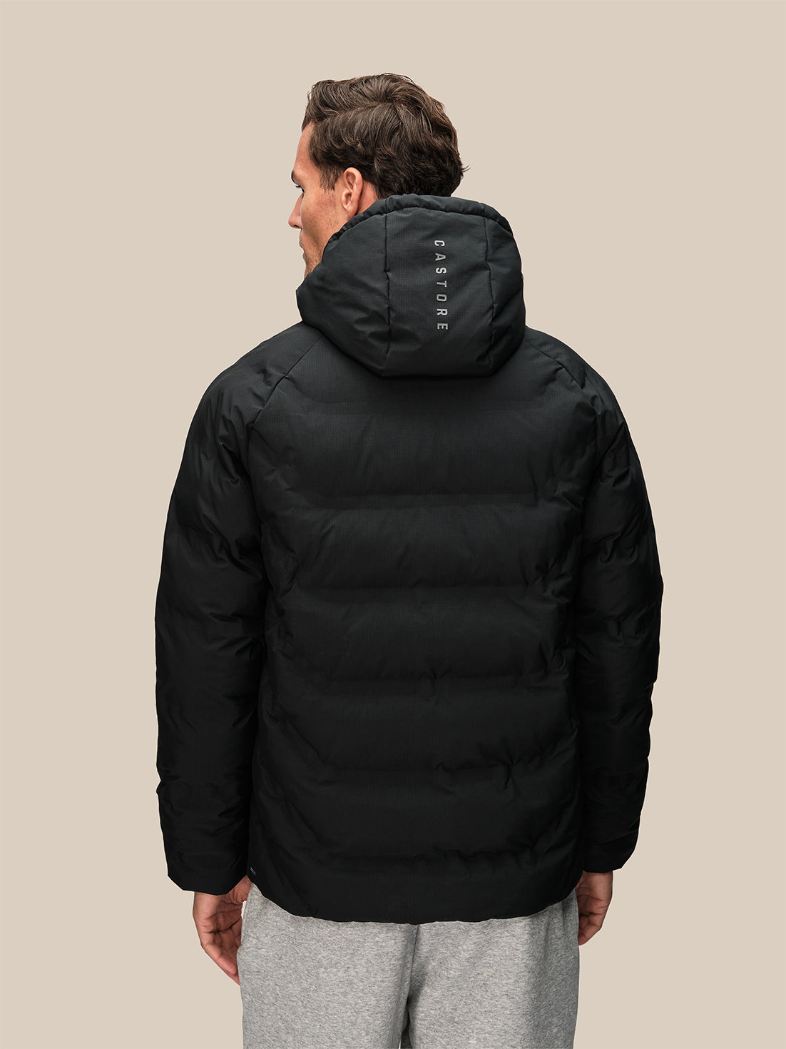 Back view of a man in a black padded hooded jacket with hood up and Castore branding on the hood