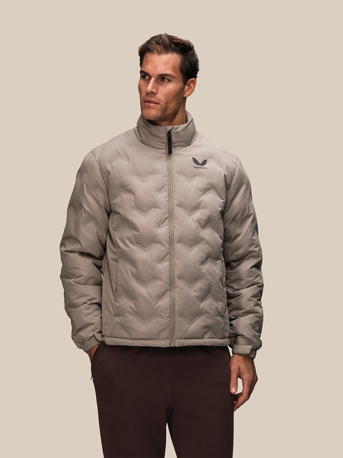 Man wearing a taupe padded quilted jacket with a stand up collar and Castore logo on the chest.