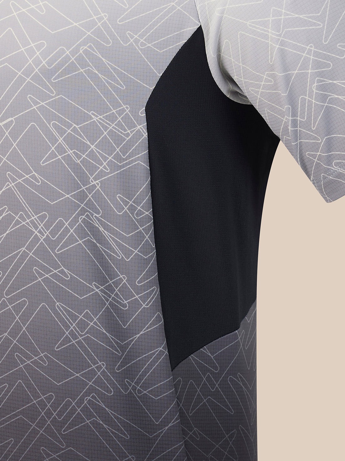 Close-up of a short-sleeve athletic jersey showing a color-blocked design with a light gray and white geometric line pattern on the upper body and sleeve, a solid black central panel, and a beige background visible on the right.