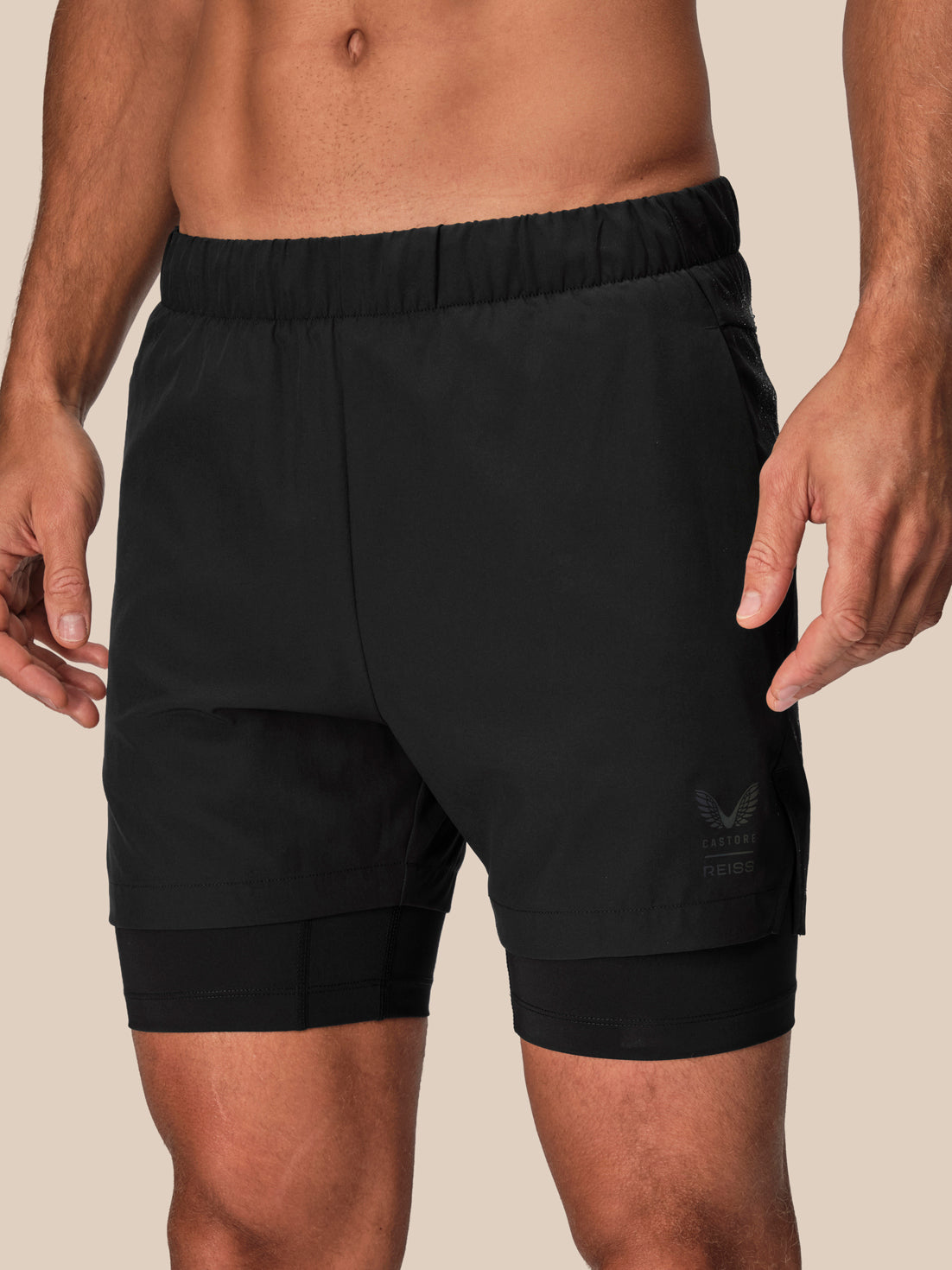 Black athletic compression shorts with a built-in second pair of legs and an elastic waistband, shown on a male model against a solid beige background.