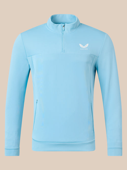 A light blue long-sleeve quarter-zip pullover jacket with a small white wing logo and the brand name 'castore' on the left chest, displayed on a solid beige background.